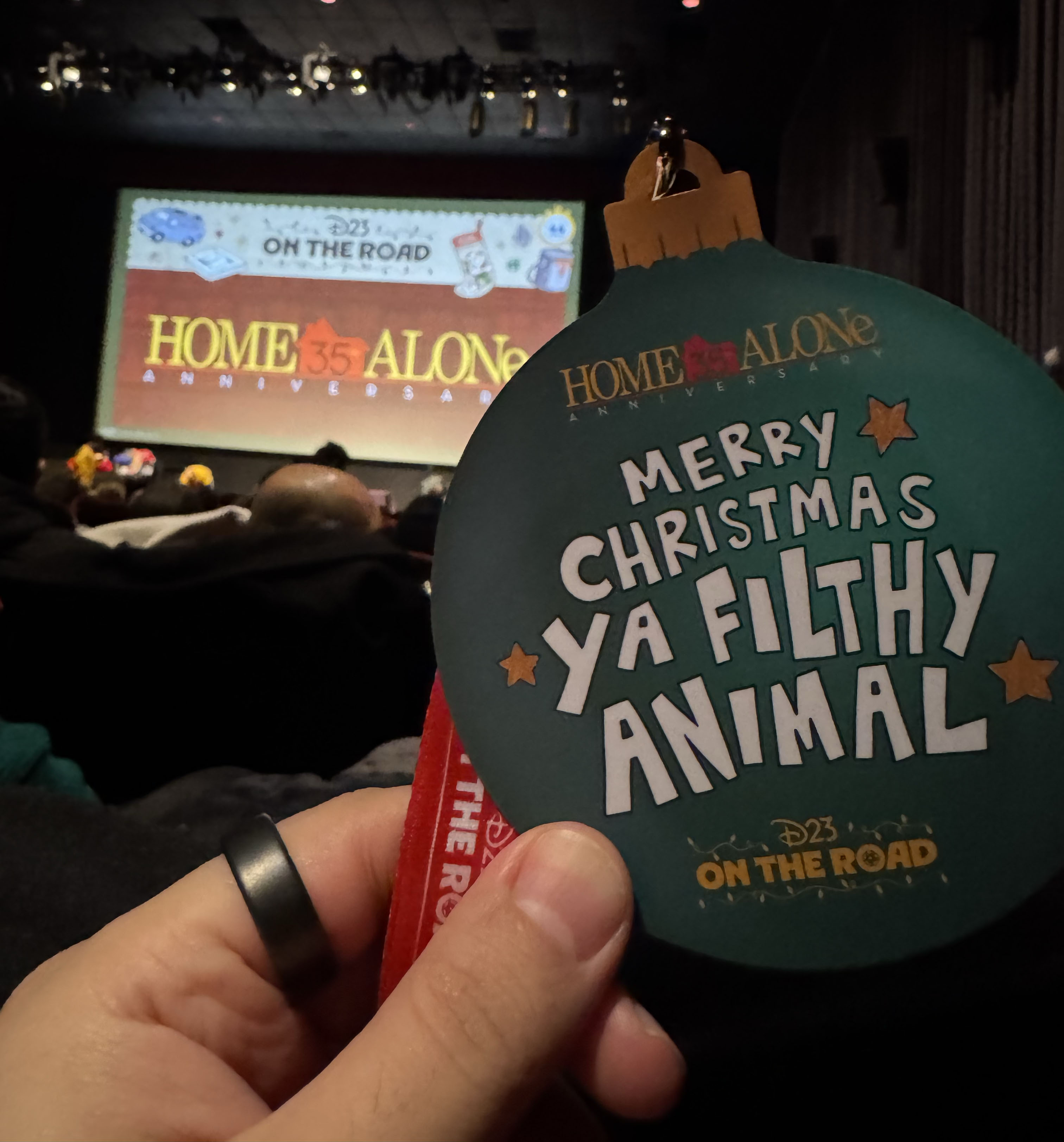 A hand holds up a Christmas ornament-shaped badge that reads “Merry Christmas ya filthy animal” with a movie screen in the background featuring the Home Alone key art.
